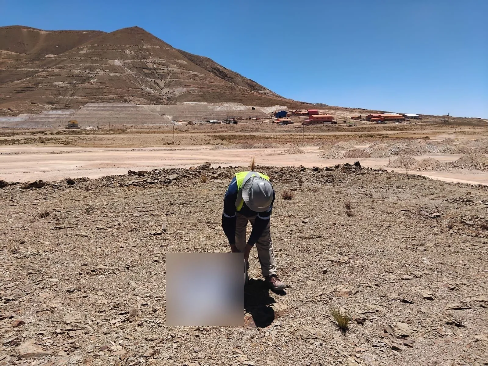 Técnico realizando un ensayo en un terreno pedregoso, con instalaciones mineras y un cerro al fondo.